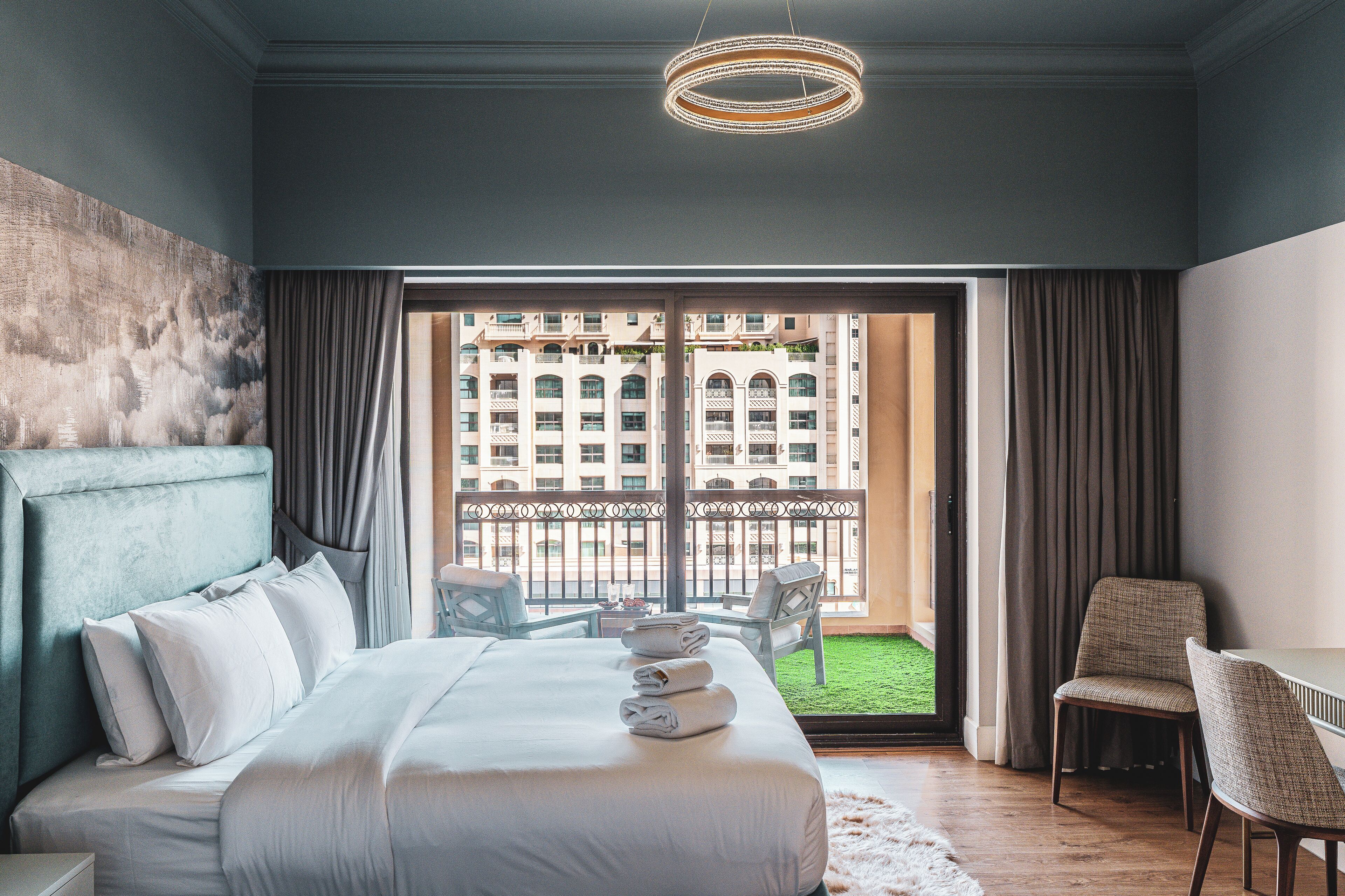 Una habitación de hotel moderna con una cama grande, vista a un edificio desde el balcón y un área de comedor con sillas.