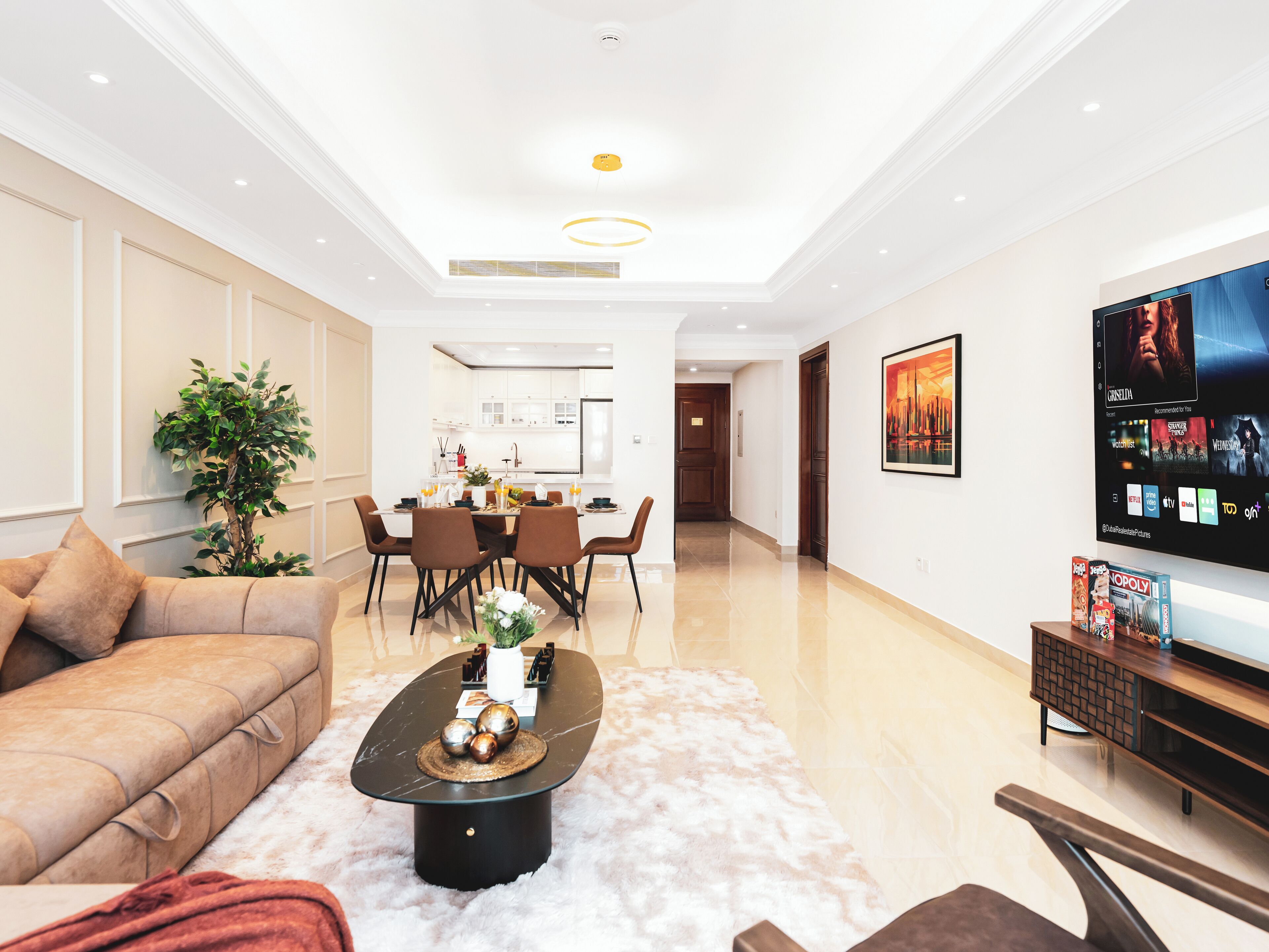 Una sala de estar moderna con sofá, mesa de centro y televisor. Se aprecia un comedor con sillas y una mesa al fondo.