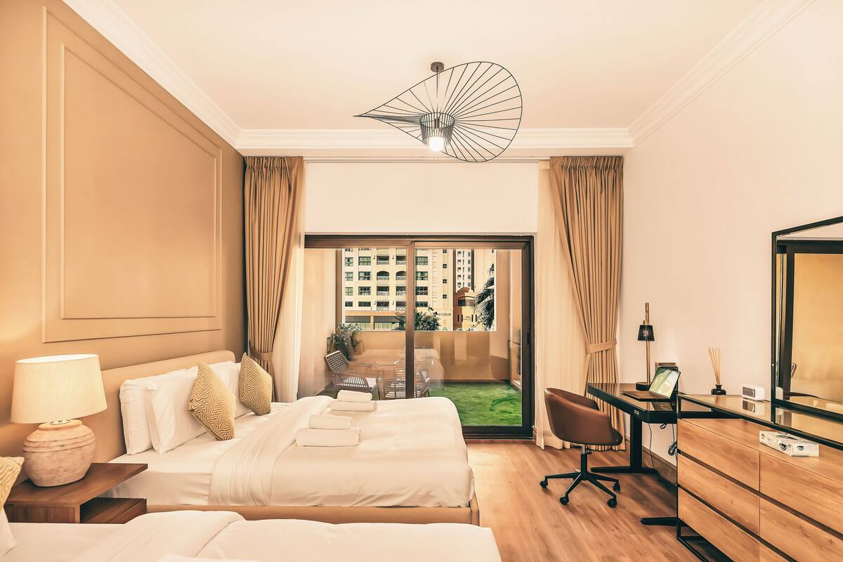 Een hotelkamer met twee bedden, een bureau, een stoel, een lamp en een groot raam met uitzicht op gebouwen.