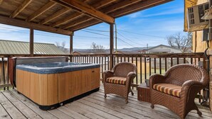 Una terrazza in legno con vasca idromassaggio, sedie in vimini e una zona riparata da cui si vedono case e montagne.