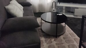 Uma sala de estar com um sofá cinzento, uma mesa de centro redonda, uma televisão de tela plana e um relógio de parede.
