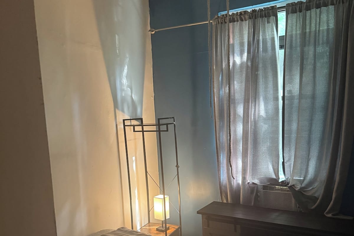 A bedroom with a bed, a bedside table with a lamp, a window with curtains, and a ceiling fan.