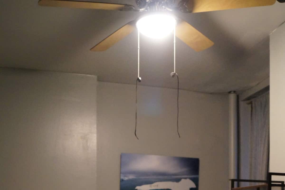 A bedroom with a single bed, a ceiling fan, a painting of a landscape, and a window with curtains.