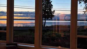 Ein Zimmer mit großen Fenstern, das einen Blick auf einen Sonnenuntergang über Wasser, einen Baum und eine Brücke bietet.