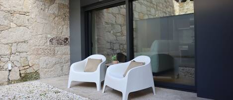 A modern outdoor seating area with two white chairs and a small table, set against a stone wall and glass door.