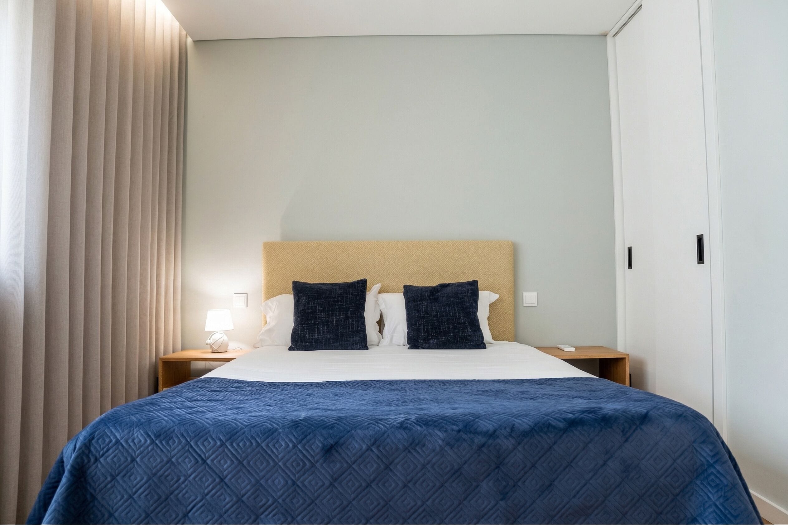 A hotel room with a bed, two pillows, a nightstand with a lamp, and a window with curtains.