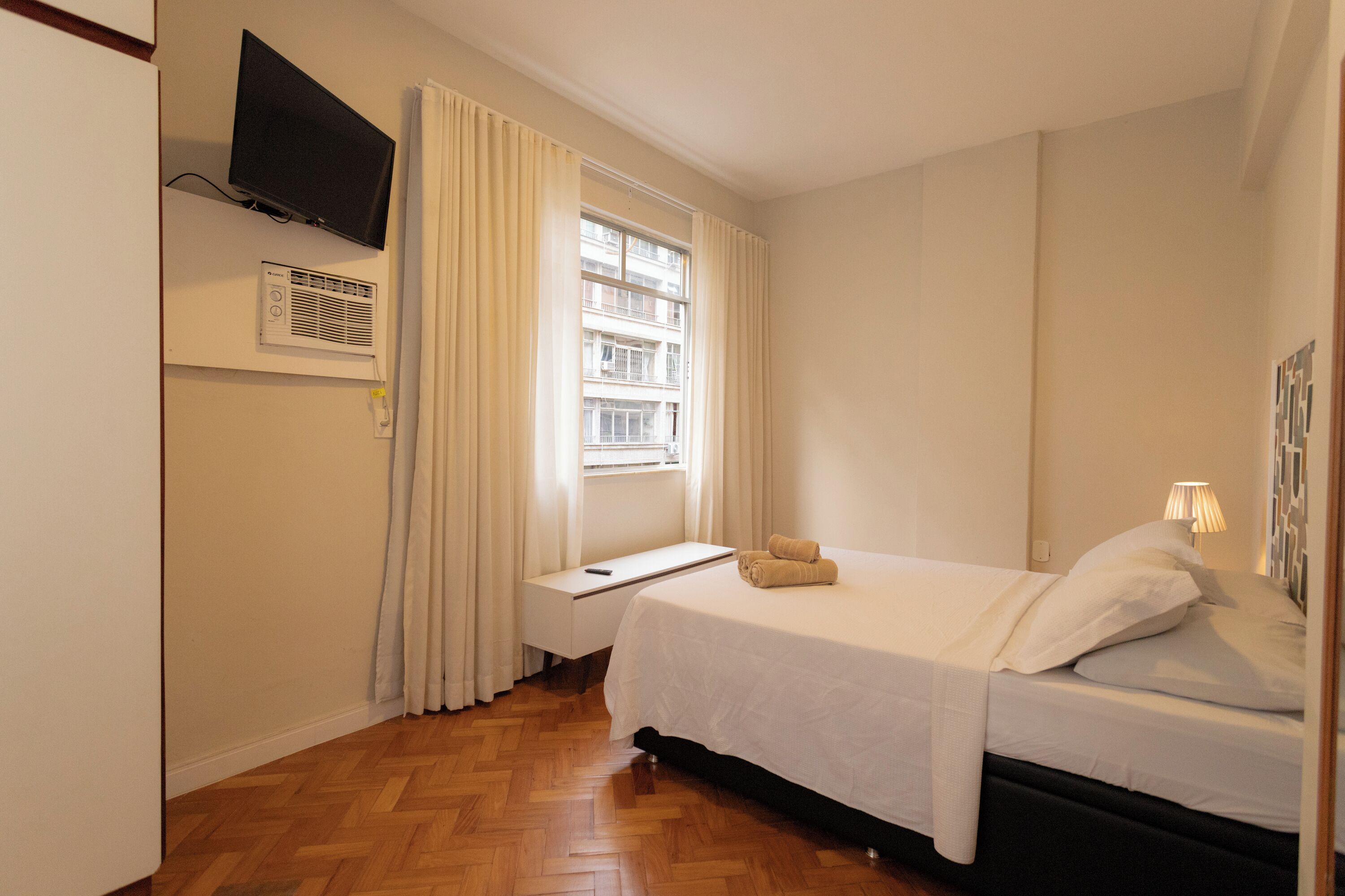 A hotel room with a bed, a TV mounted on the wall, an air conditioning unit, a window with curtains, and a bedside table.