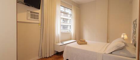 A hotel room with a bed, a TV mounted on the wall, an air conditioning unit, a window with curtains, and a bedside table.