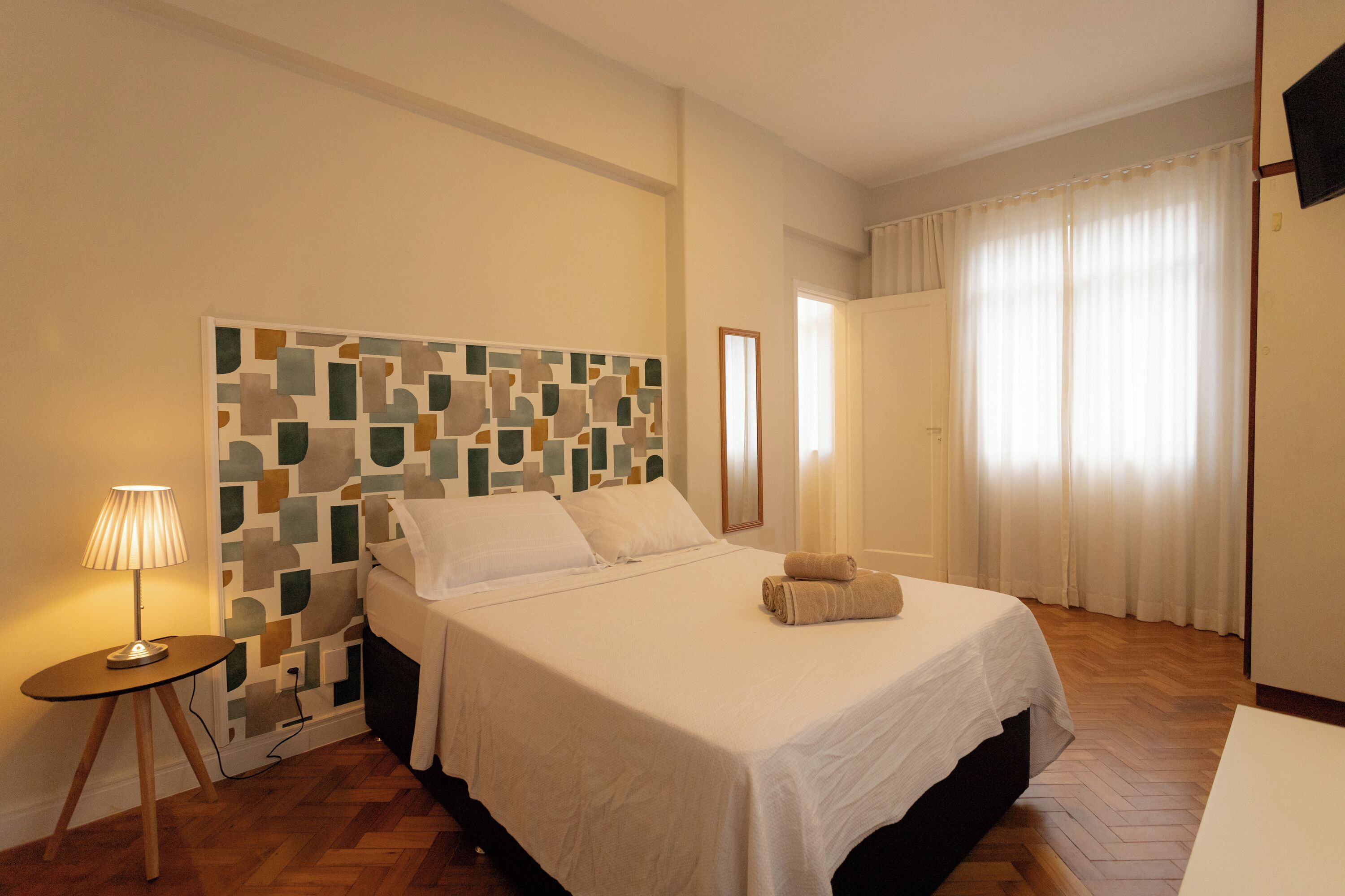 Ein ordentlich eingerichtetes Hotelzimmer mit Bett, Nachttischlampe, Wandbild und einem Fenster mit Vorhängen.