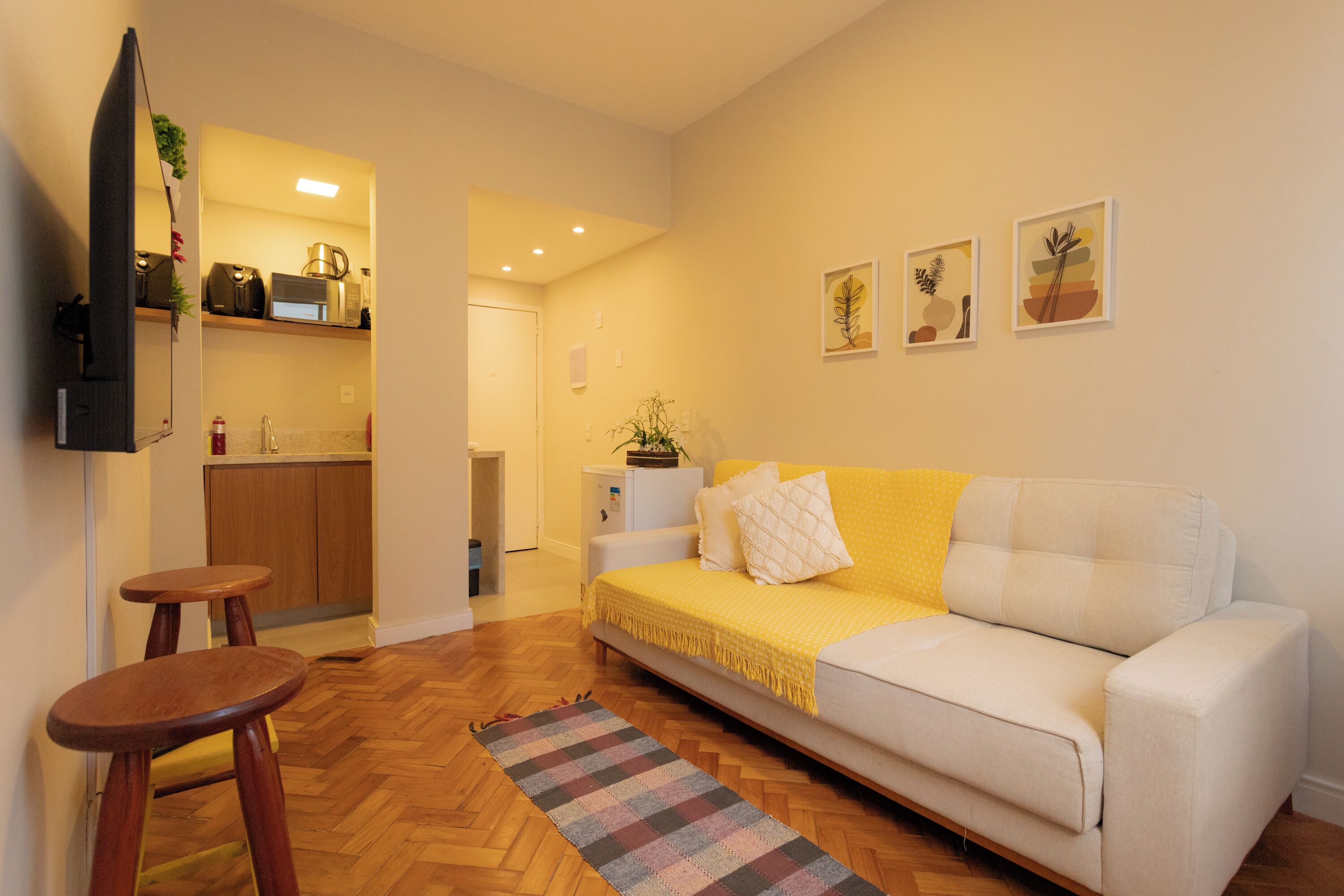 Una sala de estar con sofá, mesa de centro, piso de madera, área de cocina con armarios y electrodomésticos, y un pasillo que conduce a otra habitación.