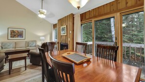 Una mesa de comedor de madera con sillas, un sofá y una chimenea en una habitación con ventanas amplias y vistas a árboles.