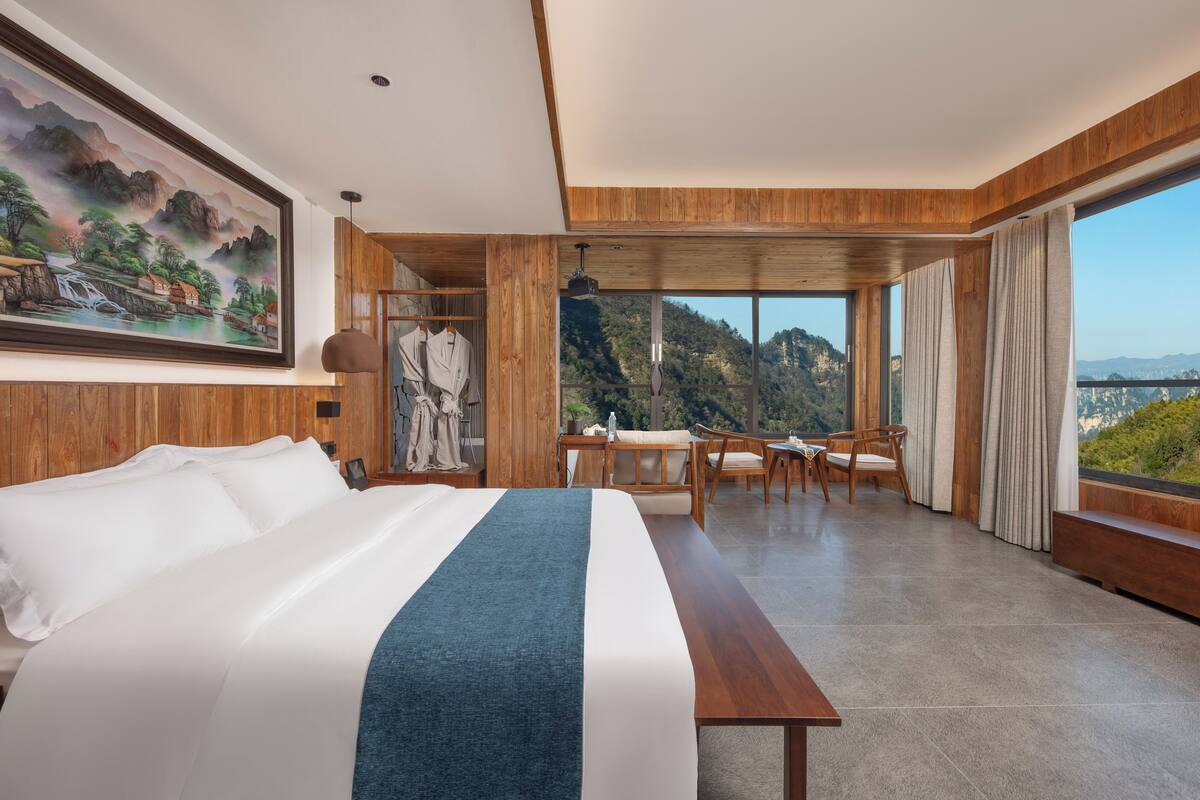 A spacious hotel room with a large bed, a dining area with a view, and a painting on the wall.
