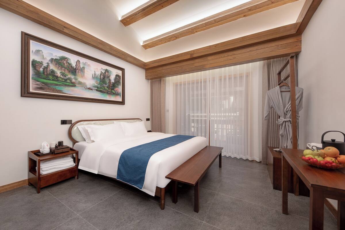 A hotel room with a bed, a wooden bench, a dining table with fruits, a painting on the wall, and a window with curtains.