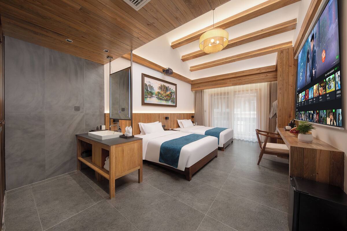 A modern hotel room with two beds, a wooden ceiling, a large TV, and a bathroom with a sink and mirror.