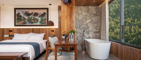 A modern bedroom with a bed, a wooden table, a white bathtub, and a large window with a forest view.