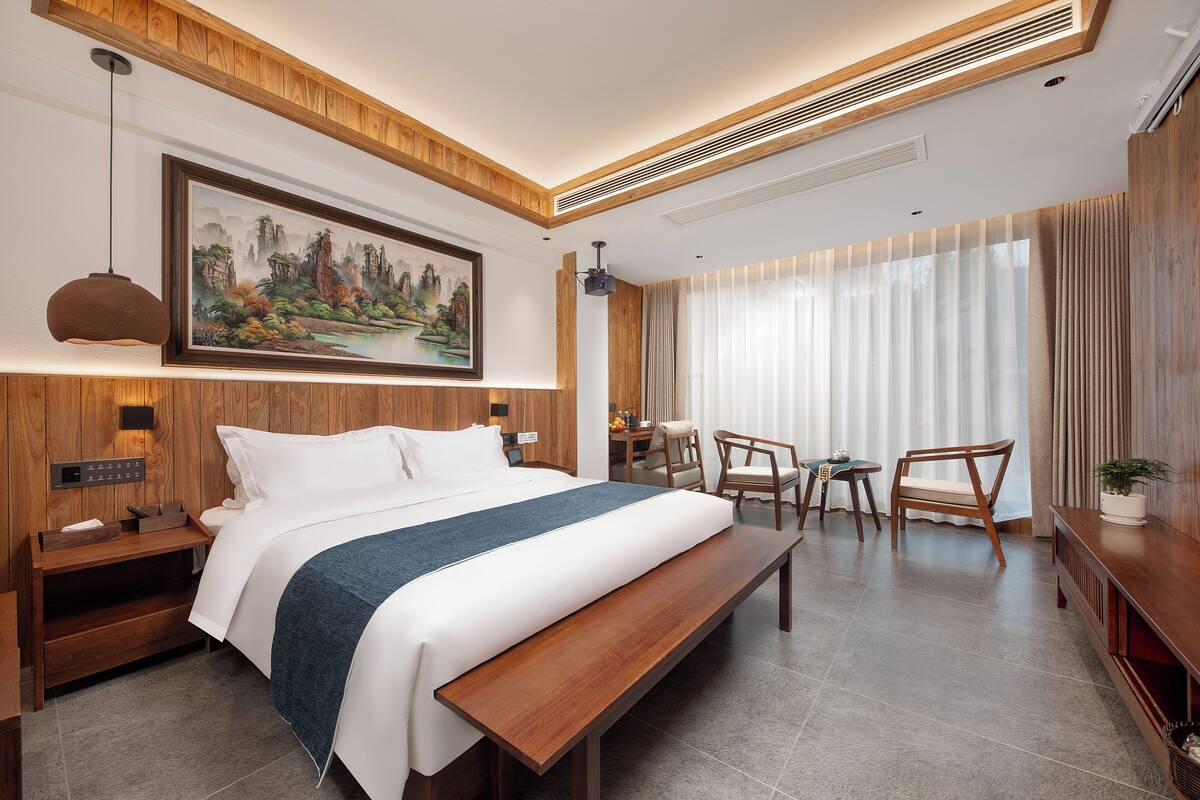 A modern hotel room with a large bed, wooden headboard, a painting, a desk, chairs, and a large window with curtains.