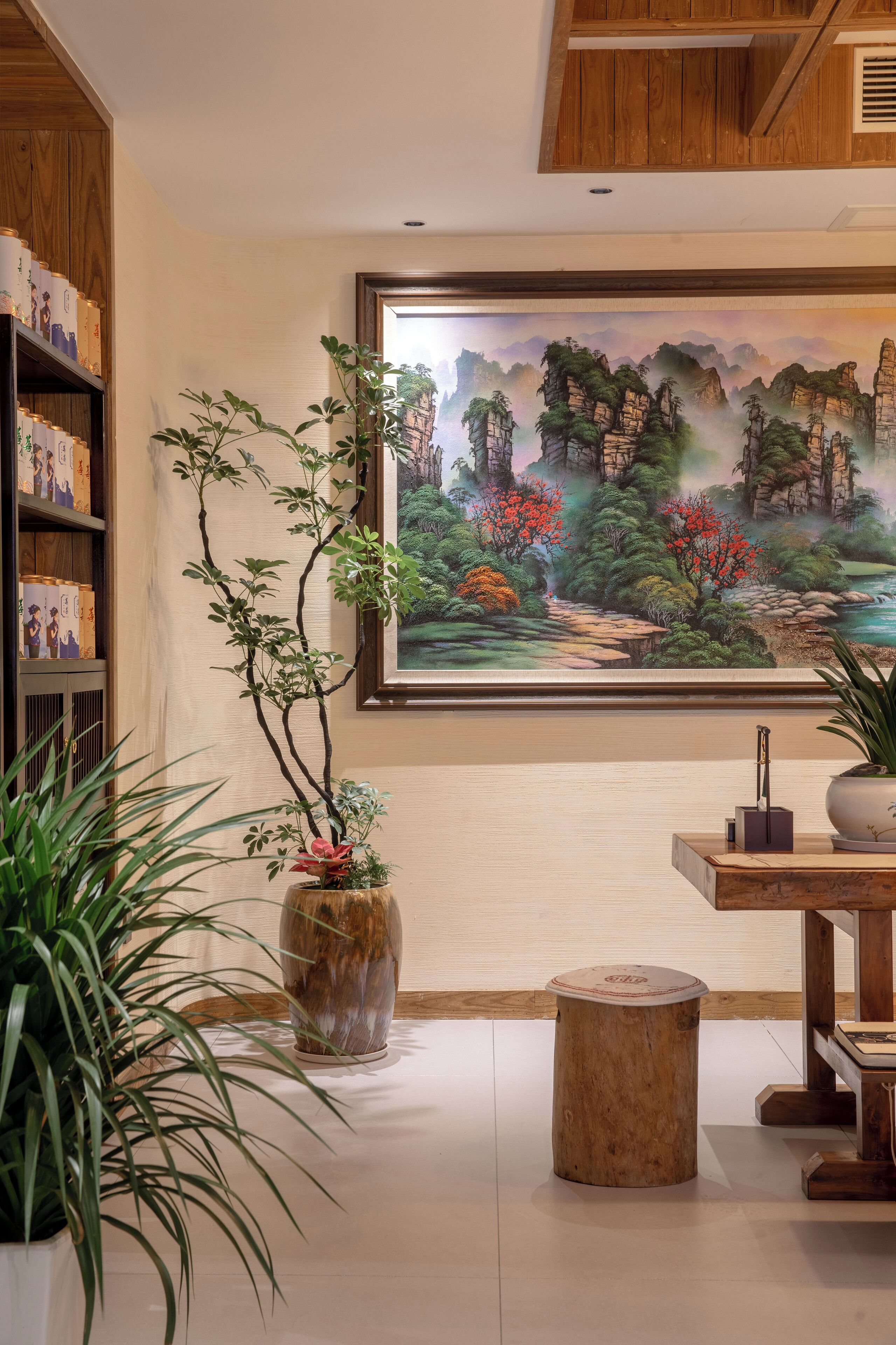 A room with a wooden bookshelf, a large painting of a landscape, a potted plant, and a round wooden stool.