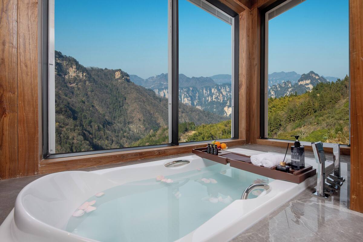 A large bathtub with a wooden tray holding towels and toiletries, set against a backdrop of a mountainous landscape through large windows.