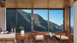 Ein Zimmer mit Holzboden, großen Fenstern, einem Esstisch mit Stühlen, einer kleinen Topfpflanze und Blick auf eine gebirgige Landschaft.