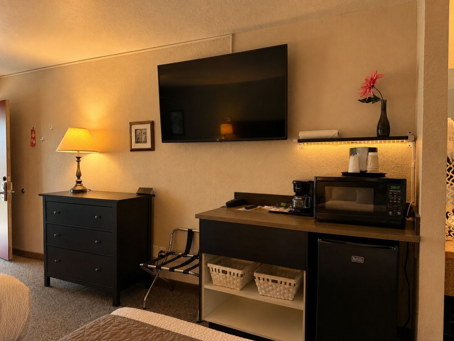 A hotel room with a flat-screen TV, a coffee maker, a microwave, a mini-fridge, a desk with a lamp, a chair, and a bed.