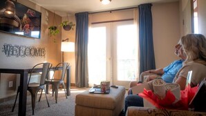 A living room with a sofa, a coffee table, and a TV mounted on the wall. There is a window with curtains, a small table with chairs, and a welcome sign on the wall. - Mountain View Lodge and Resort (Manson)
