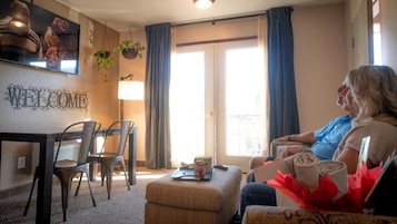 Una sala de estar con un sofá, una mesa de centro y un televisor montado en la pared. Se observa una ventana con cortinas, una pequeña mesa con sillas y un letrero de bienvenida en la pared.