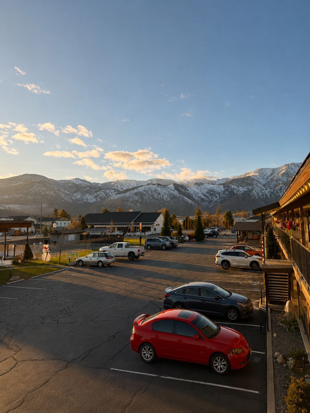 Un estacionamiento con varios autos, un edificio con porche y montañas nevadas al fondo.