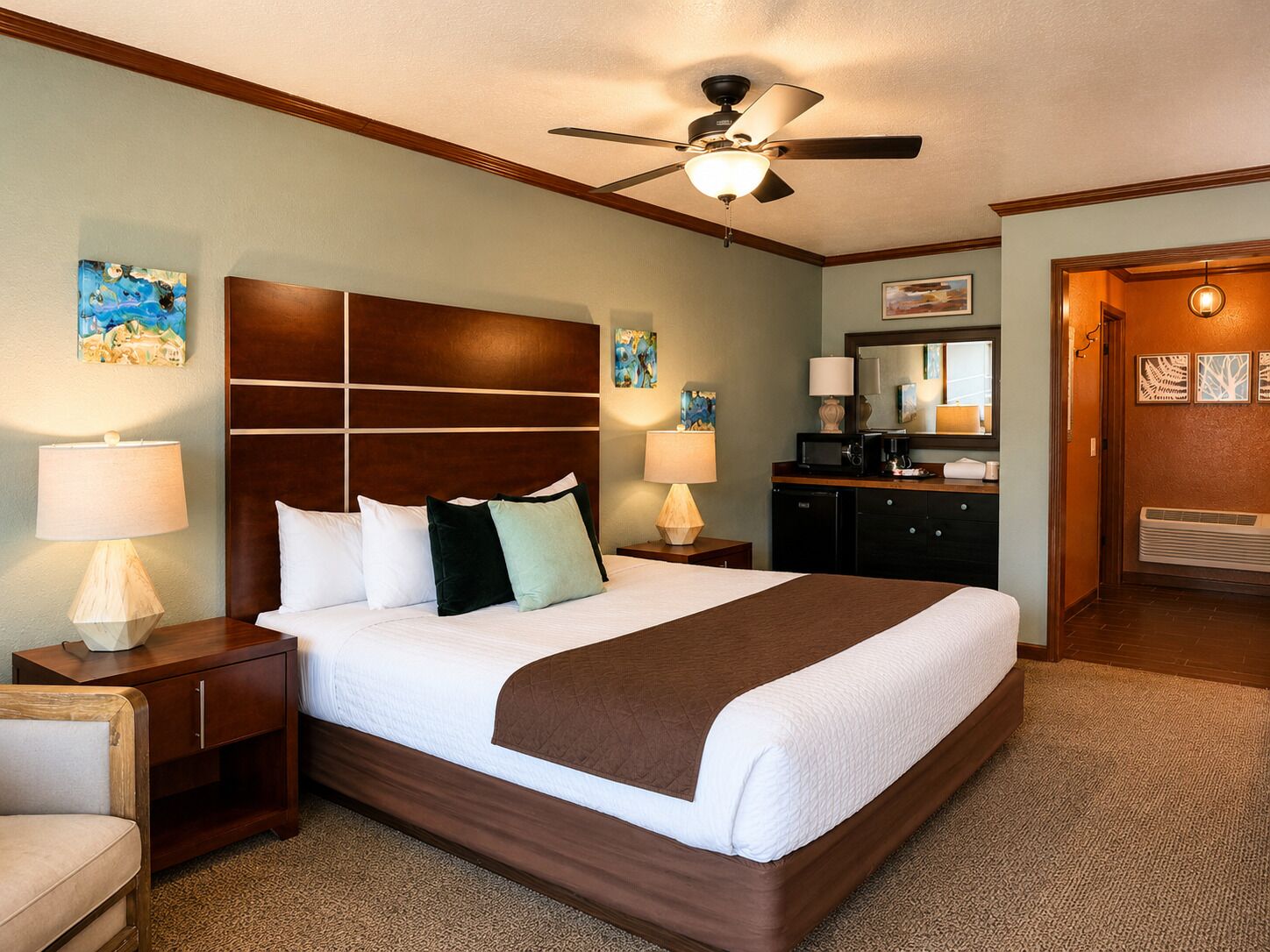 Quarto de hotel com uma cama grande, luminárias de cabeceira, ventoinha no teto, obras de arte nas paredes e cômoda com espelho.