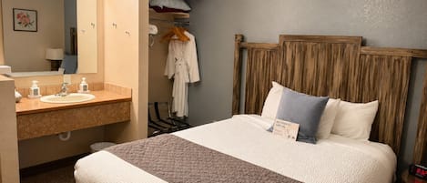A hotel room with a bed, a bedside table, a nightstand, a mirror, a vanity with a sink, and a robe hanging on a rack.