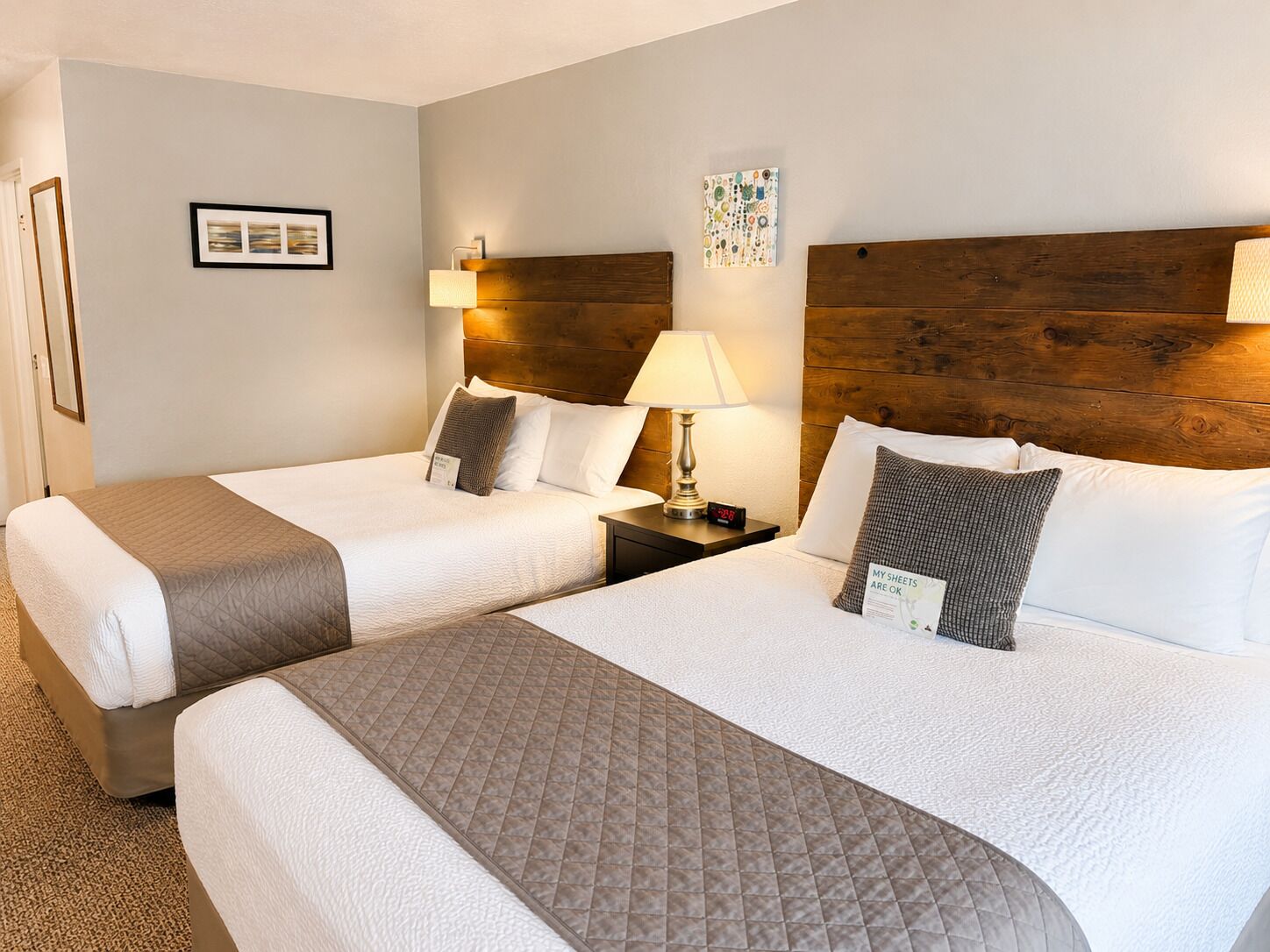 A hotel room with two beds, a wooden headboard, a nightstand with a lamp, and a wall clock.