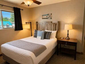 A neatly made bed with pillows, a bedside table with a lamp, a ceiling fan, and a wall sign with directional arrows. - Mountain View Lodge and Resort (Manson)