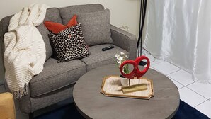 Un salón con un sofá gris, una mesa de centro redonda, un cuadro en la pared, una lámpara de pie y una cortina blanca.