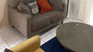 Un salón con un sofá gris, una mesa redonda de madera, una alfombra azul, un cuadro en la pared y un ventilador de techo.