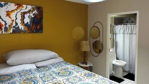 Un dormitorio con una cama, un cuadro, una lámpara y un baño visible a través de una puerta abierta.