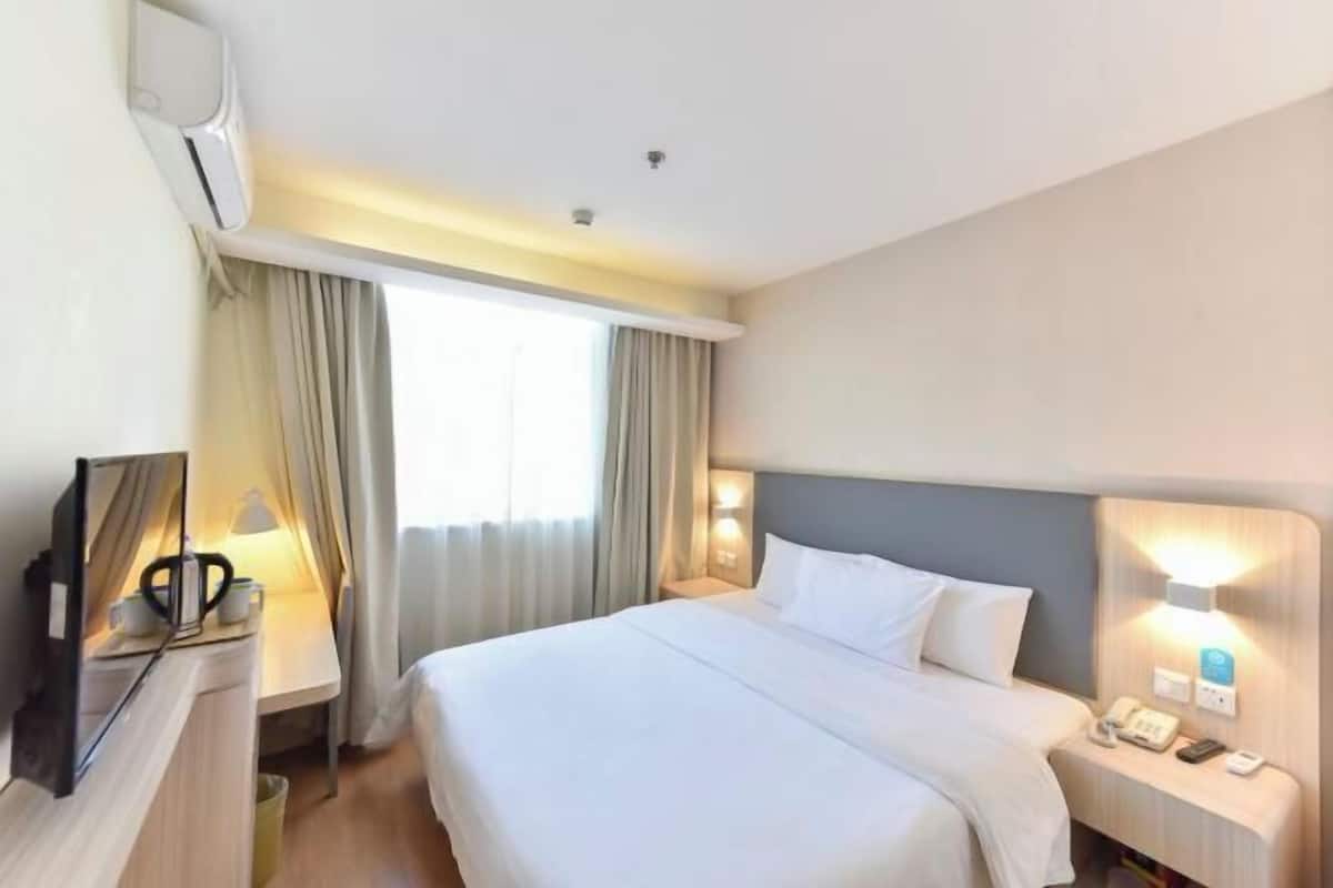Habitación de hotel con cama, mesita de noche con lámpara, escritorio con computadora y unidad de aire acondicionado.