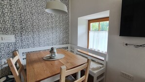 Una habitación con una mesa de comedor de madera, un banco blanco, una ventana con vistas a árboles y un televisor montado.