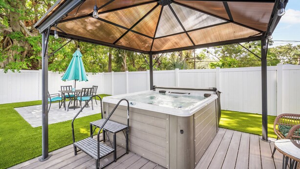 Ein Whirlpool unter einer Markise mit einem Patio-Tisch und Stühlen, umgeben von einer Holzterrasse und einem weißen Zaun.