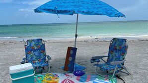 Ein Strandaufbau mit zwei Liegestühlen, einem blauen Sonnenschirm, einer Kühlbox, einem Strandtuch und einer Strandtasche.