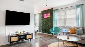Ein modernes Wohnzimmer mit Flachbildfernseher, einer Couch, einem Couchtisch und einem an der Wand montierten Fernseher. Eine grüne Wand mit einem Neonzeichen, ein Fenster mit Jalousien und ein Deckenventilator sind ebenfalls vorhanden.