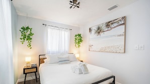 Ein ordentlich eingerichtetes Schlafzimmer mit Bett, Nachttischlampen, einem Wandbild und hängenden Pflanzen.
