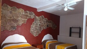 Une chambre avec deux lits, un mur en pierre, un ventilateur de plafond et un téléviseur fixé au mur.