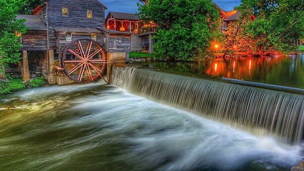 A historic watermill with a large wheel, a wooden building with a porch, and a river.