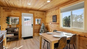 Puinen mökki, jonka sisustuksessa on ruokapöytä, tuoleja, joulukuusi ja kirjahylly.