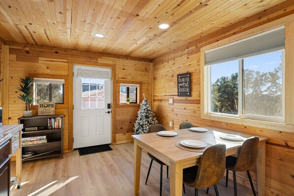 A wooden cabin interior with a dining table, chairs, a Christmas tree, and a bookshelf.