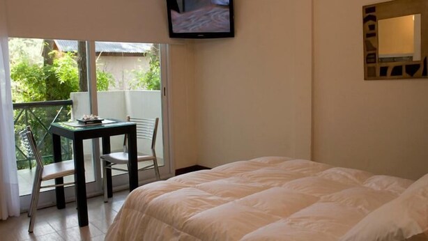 Ein Schlafzimmer mit einem Bett, einem an der Wand montierten Fernseher, einem Tisch mit Stühlen und einer Glastür zu einem Balkon.