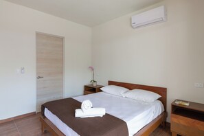 A neatly made bed with a brown blanket and a rolled towel, a wooden nightstand with a book, a small potted plant, and an air conditioning unit on the wall.