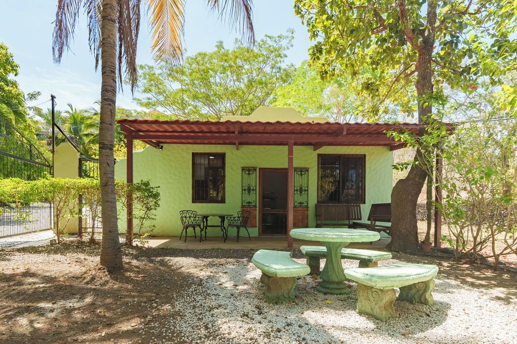 A small building with a green exterior, a covered patio with a table and chairs, and a gravel ground.