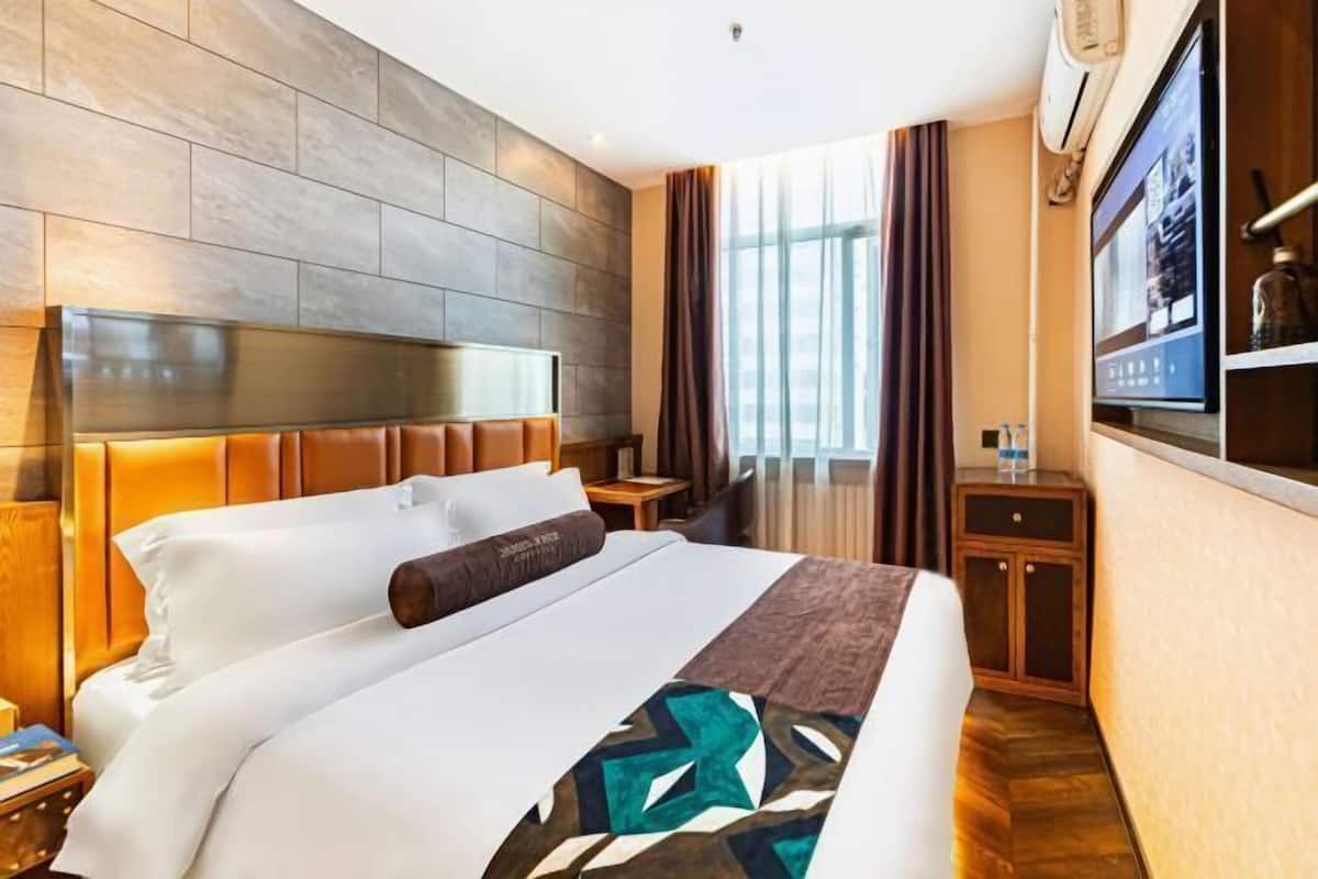 Ein Hotelzimmer mit einem großen Bett, einem Kopfteil, einem Nachttisch, einem Fernseher und einem Fenster mit Vorhängen.