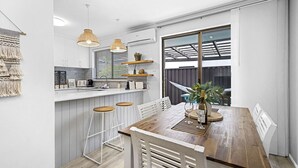A modern kitchen with white cabinetry, a wooden dining table, and bar stools. There are pendant lights, a window with blinds, and a plant on the table. - Coastal convenience - 2 min walk to shops & cafes (Scarborough)
