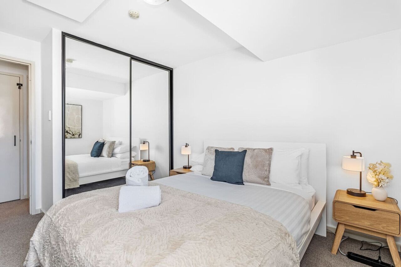 A modern hotel room with a large bed, a bedside table with a lamp, a mirror, and a wardrobe.